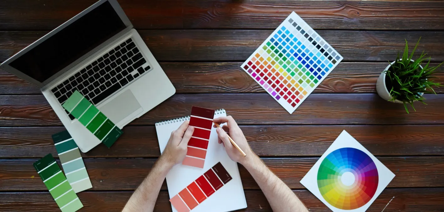 Top-down view of a design desk with red and green paint swatches, a color chart, a color wheel and a laptop